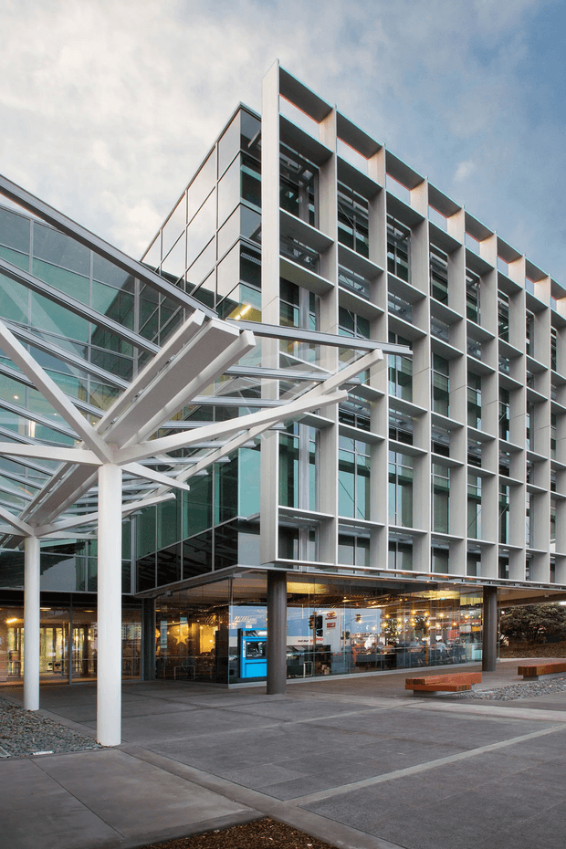 Exterior architectural photograph of a contemporary multi-storey commercial building. The facade features a strong repetitive grid of projecting white concrete or metal fins, extensive glazing, recessed balconies or openings, and a transparent glazed lower-level entrance or retail frontage. A dramatic white steel-and-glass canopy projects over the paved forecourt, with built-in timber benches and dark columns. The composition is photographed from a low angle beneath a cloudy sky.