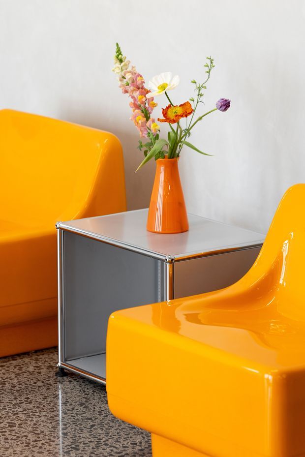 A close-up interior vignette featuring two bright orange, glossy molded lounge chairs around a square side table with a reflective metal frame and pale top. An orange vase holding a small colorful flower arrangement sits on the table. The space has a plain light wall and a speckled terrazzo-style floor, creating a bold, playful mid-century-inspired composition.