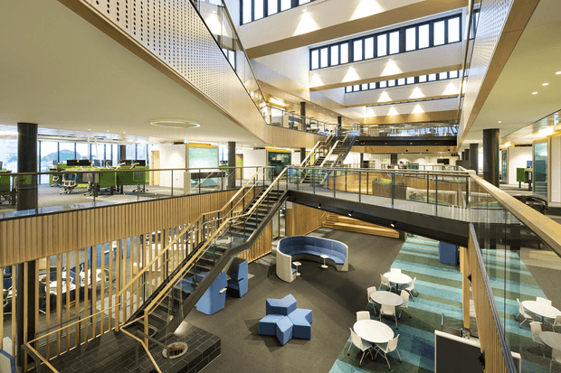 A large contemporary multi-level commercial interior with an expansive atrium, multiple staircases, bridges, glass balustrades, timber-lined walls, exposed structural elements, and clerestory glazing. Informal lounge seating and café-style tables occupy the lower level, while open work areas are visible around the perimeter. The space is bright, airy, and highly connected vertically.