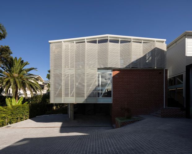 Exterior view of a contemporary multi-level building with a projecting upper volume wrapped in fine horizontal slatted screening, contrasting dark red brick walls, extensive glazing, and a flat roof. The building sits above a paved forecourt with palms and hedging visible at the side.