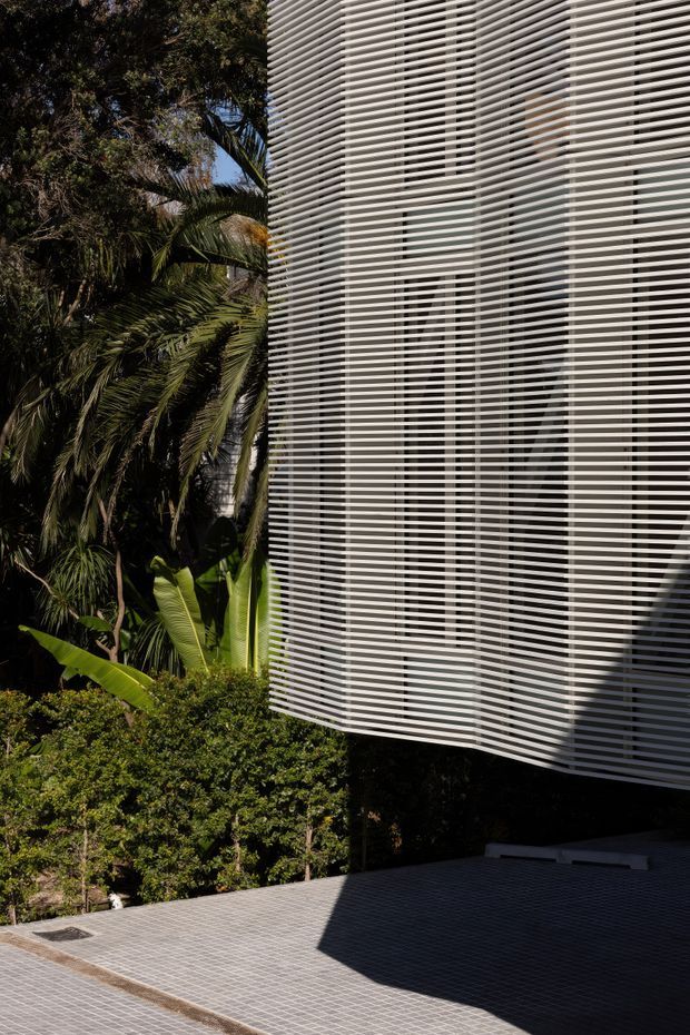 Exterior architectural photograph showing a contemporary building facade screened by dense horizontal white louvres. Glazed openings and shadowed interior spaces are visible behind the screen. The building projects above a paved forecourt beside lush tropical landscaping with palms and broad-leaf plants. Strong sunlight creates deep shadows across the grey tiled paving.