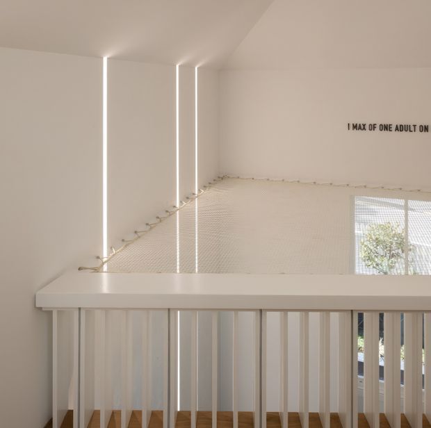 Minimalist white stair landing with a white-painted balustrade, timber stair treads, slim vertical light slots, and safety netting stretched across the upper stair void. A partial occupancy notice is visible on the wall, with daylight and greenery seen through the window.