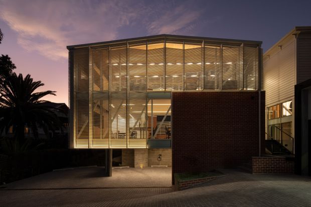 Twilight exterior view of a contemporary multi-level building with a large illuminated glazed volume screened by horizontal louvres, a dark brick-clad lower section, and an adjacent connected structure. The facade opens onto a paved driveway or forecourt, with warm interior lighting visible through the glazing.