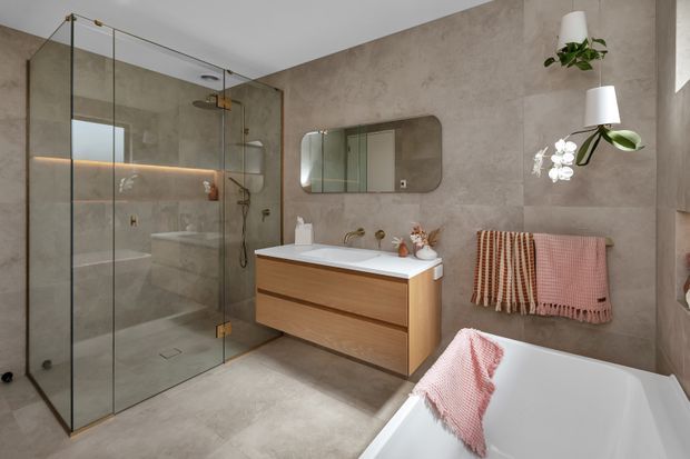 Contemporary bathroom featuring a frameless glass walk-in shower, wall-mounted timber vanity with white basin, freestanding white bathtub, large-format warm grey stone-look wall and floor finishes, integrated mirror, and soft pink and terracotta textiles. Brass fixtures and subtle wall lighting add warmth.