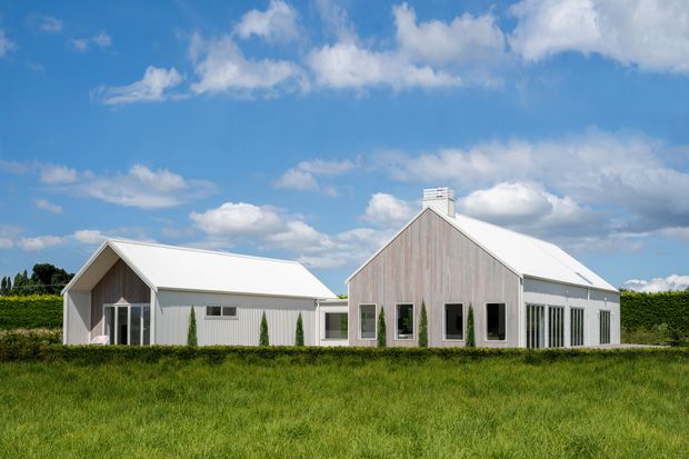A low-slung contemporary rural property composed of connected gabled volumes. The buildings feature pale vertical timber-like cladding, crisp white metal pitched roofs, dark-framed windows, and a broad grassy foreground beneath a bright blue sky.