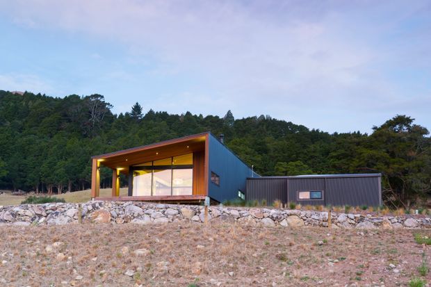 A low, contemporary single-storey house set against a wooded hillside. The compact building has a dark metal-clad rectangular wing, a warm timber-lined recessed entrance or living area, extensive glazing, and a broad shallow roof overhang. A separate dark-clad outbuilding sits to the right, while a rocky retaining edge and dry ground occupy the foreground.
