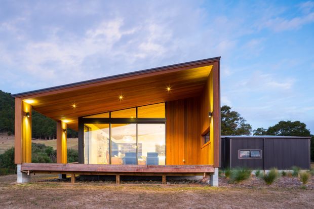 Exterior view of a compact modern timber-clad pavilion or cabin with a dramatic asymmetrical sloping roof and deep covered outdoor terrace. Warm timber soffits and columns frame full-height glazing, while a darker low ancillary volume sits to the side. The building is set in a sparse landscaped rural or semi-rural site and photographed at dusk with interior lighting glowing through the glass.