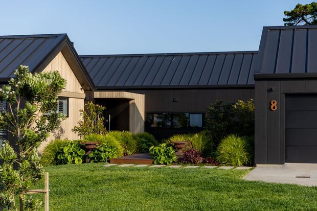 Exterior view of a contemporary single-storey home with dark standing-seam metal roofing, dark cladding, timber-toned gabled entry volume, and a prominent integrated garage. A landscaped front garden with layered planting and lawn leads to the recessed entrance. The composition shows a connected series of pitched and low roof forms with clean modern detailing.