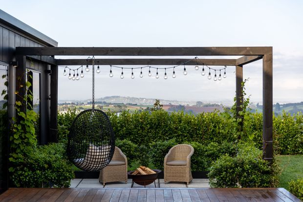 An outdoor terrace overlooking a broad rural or suburban landscape. A dark timber pergola frames the seating area, with string lights overhead, a hanging woven egg chair, two wicker armchairs, and a small fire bowl. Dense hedging and climbing greenery provide privacy, while timber decking forms the foreground.