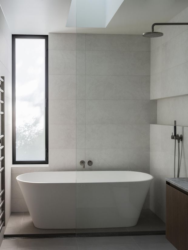 Modern minimalist bathroom featuring a freestanding white bathtub beneath a large skylight, full-height light grey tiled walls, a frameless glass shower enclosure, matte black shower fittings, and a tall black-framed frosted window. A dark wood vanity with a grey stone benchtop is partially visible at right, while a heated towel rail appears at left.