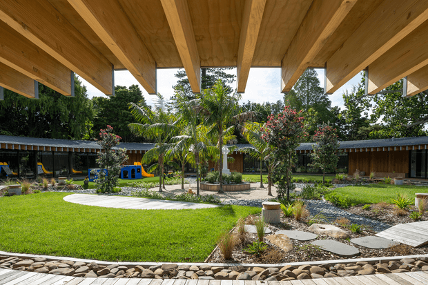 A landscaped courtyard viewed from beneath a broad timber-beamed roof overhang. The space features lush lawn, tropical planting, paved and stepping-stone paths, a shallow rock-lined water feature or drainage channel, raised timber decking, and low pavilion-like buildings around the courtyard. Children's play equipment is visible in the background.