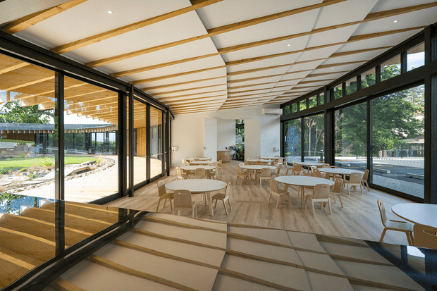 A bright contemporary dining and café-style interior with extensive black-framed floor-to-ceiling glazing on both sides, overlooking landscaped outdoor areas. The space features light timber-look flooring, white walls, exposed timber ceiling battens with white acoustic ceiling panels, and numerous round light-wood tables with matching chairs. A stepped transition and reflective glazing add visual depth, while abundant daylight creates an airy, open atmosphere.