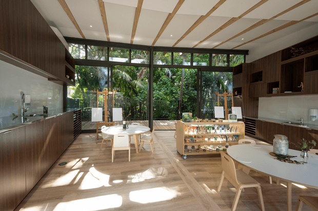 A bright, open-plan interior with a kitchen and dining area, featuring dark timber cabinetry, a pale stone or solid-surface benchtop, large black-framed glazed doors and windows opening toward lush greenery, and a white ceiling with exposed timber battens. Two light timber dining tables and chairs sit around a central display island, creating a refined, nature-connected space.