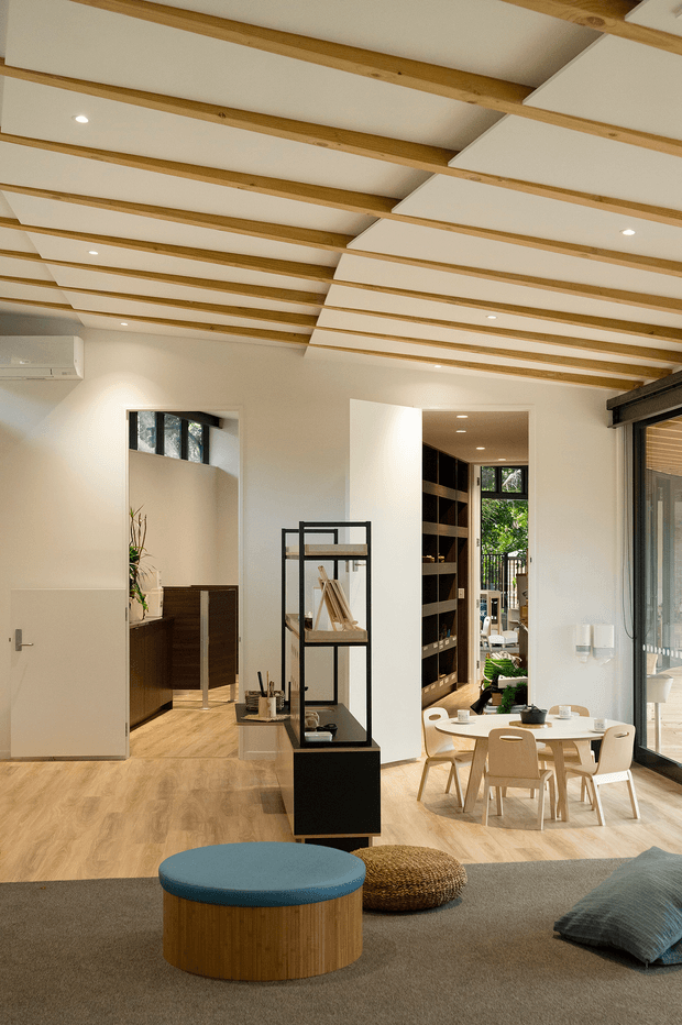 A bright, open-plan contemporary interior combining a living room, dining area and visible kitchen zone. The room features a warm timber floor, white walls, exposed timber ceiling beams with white acoustic or recessed panels, extensive glazing, black-framed built-in shelving, and light timber dining furniture. A round blue upholstered coffee table and textured floor cushions add restrained colour to the neutral scheme.