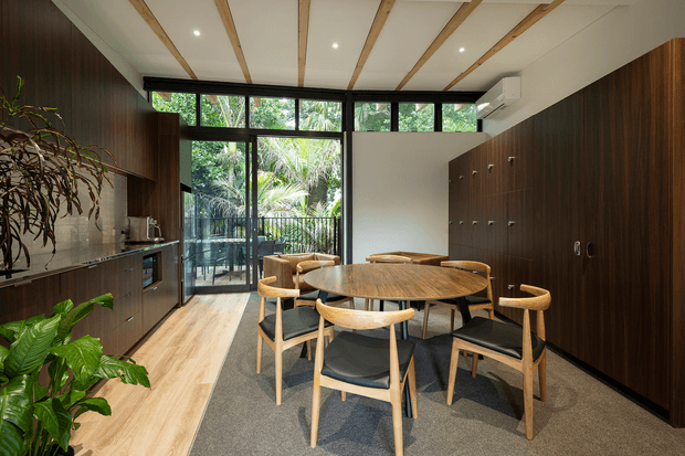 A refined open-plan kitchen and dining area with dark timber cabinetry, a stone benchtop, light timber flooring, and a round timber dining table surrounded by timber-framed chairs with black seats. Full-height glazing and a glazed door open to lush greenery, while exposed timber ceiling battens, recessed lighting, and dark-framed windows add architectural character. Indoor plants bring an organic element to the warm, contemporary space.