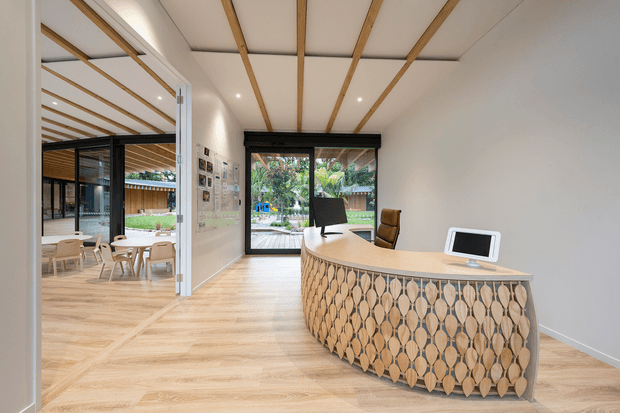 Bright contemporary reception area with a distinctive curved timber-fronted reception desk, pale timber flooring, and a white ceiling detailed with exposed light-wood battens. Full-height black-framed glazing opens views toward a landscaped outdoor area. A glazed opening to the left reveals an adjoining informal dining or breakout space with tables and chairs.