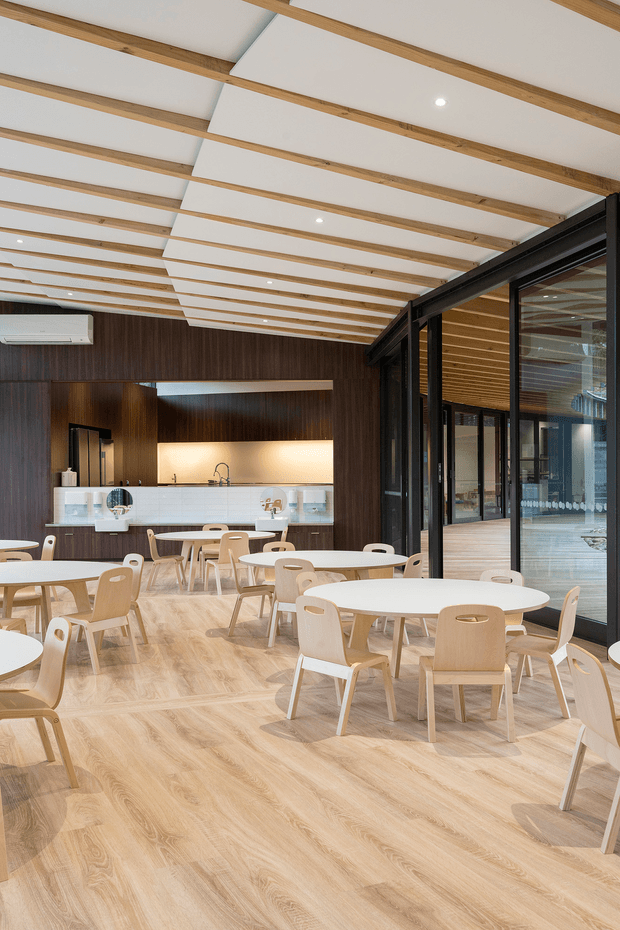 A bright contemporary dining and café-style communal space with pale timber tables and chairs arranged across a light timber-look floor. Dark timber wall panelling frames a compact kitchenette or serving counter at the rear, while black-framed glazed doors open to a timber-lined adjoining area. The ceiling features exposed light timber beams with recessed downlights, creating a warm, clean and welcoming interior.