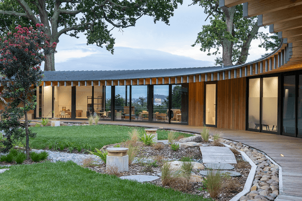 A low-profile contemporary residential building arranged around a landscaped courtyard. The long single-storey wing has dark standing-seam metal roofing, extensive floor-to-ceiling glazing, warm vertical timber cladding and exposed timber roof framing. A timber deck and gently curving stone-edged pathway lead through a naturalistic garden with lawn, ornamental grasses and established trees.