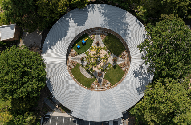 Aerial top-down view of a large circular building with a broad ribbed metal roof surrounding a landscaped central courtyard. The courtyard contains curved lawn areas, stone paths, planted garden beds, and a central sculptural planting arrangement. Dense mature trees frame the property, with a small adjacent structure and marked parking visible at the edges.