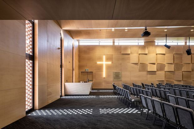 A contemporary chapel or worship auditorium with warm timber wall and ceiling lining, rows of upholstered chairs facing a simple reception or altar area, a glowing cross, patterned timber acoustic wall panels, concealed linear lighting, and ceiling-mounted audiovisual equipment. Daylight filters through perforated brick or decorative screens at the side, creating patterned shadows across the dark carpet.