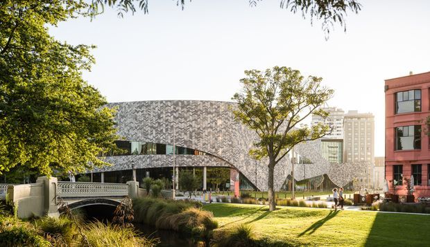 Exterior architectural view of a contemporary civic or commercial building with a curved, sculptural facade clad in a patterned grey material. The building sits within a landscaped urban setting with mature trees, lawn, ornamental grasses, pedestrian paths, and a small bridge over a planted water feature. A red masonry building and taller urban structures are visible in the background.