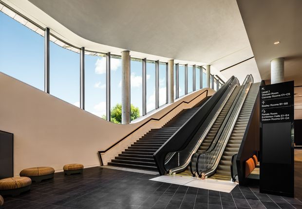 A spacious contemporary public interior featuring a broad staircase beside two escalators, framed by tall curved floor-to-ceiling windows and cylindrical structural columns. Dark tiled flooring, pale walls and ceiling, black stair and escalator finishes, and a black wayfinding directory create a refined institutional or commercial atmosphere.