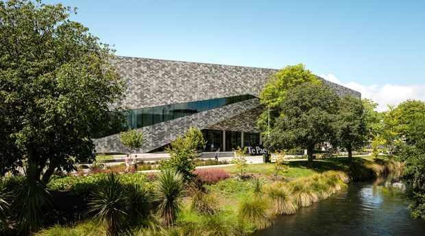 Exterior architectural photograph of a contemporary multi-level building beside a landscaped waterway. The building features a distinctive grey patterned cladding facade, angular projecting and recessed volumes, a long band of dark glazing, and a landscaped forecourt with mature trees and ornamental grasses.