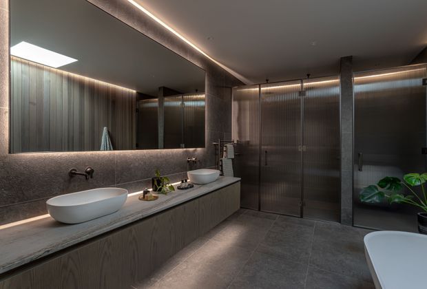 A spacious, moody contemporary bathroom with a long floating double vanity, two white vessel basins, wall-mounted brushed-metal taps, and a large illuminated mirror. The vanity features dark wood-grain cabinetry and a grey stone benchtop. Large grey floor and wall tiles, ribbed translucent glass shower enclosures with dark metal frames, recessed linear lighting, and a partially visible freestanding white bathtub create a refined, hotel-like atmosphere. A potted plant adds a small organic accent.