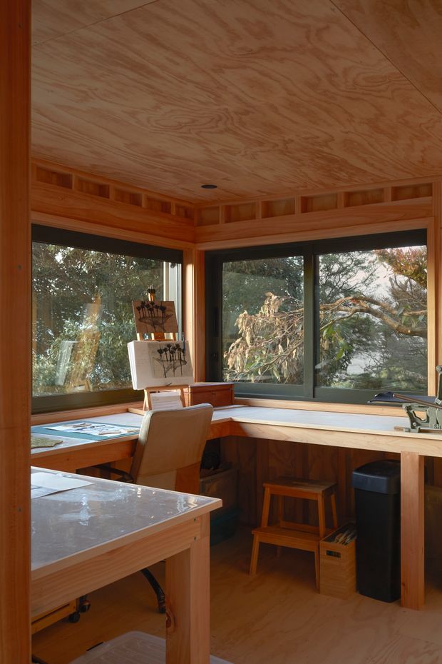 A compact, timber-lined home office or studio with extensive corner glazing overlooking trees. Built-in timber desks wrap around the windows, with a drafting table, office chair, stools, artwork, and a small dark waste bin. The ceiling, walls, cabinetry, and floor are all warm natural timber, while dark window frames provide contrast.