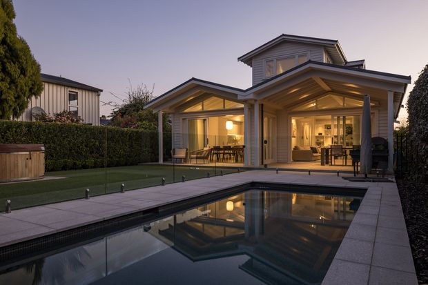 A dusk exterior view of a refined two-storey suburban home with pale weatherboard cladding, projecting gabled roof forms and warmly lit glazed living areas. A long rectangular swimming pool occupies the foreground, bordered by large-format pale paving, clipped hedges and a manicured lawn. The open-plan interior and dining area are visible through the glazing, while timber-lined soffits and exposed roof structure add warmth to the facade.