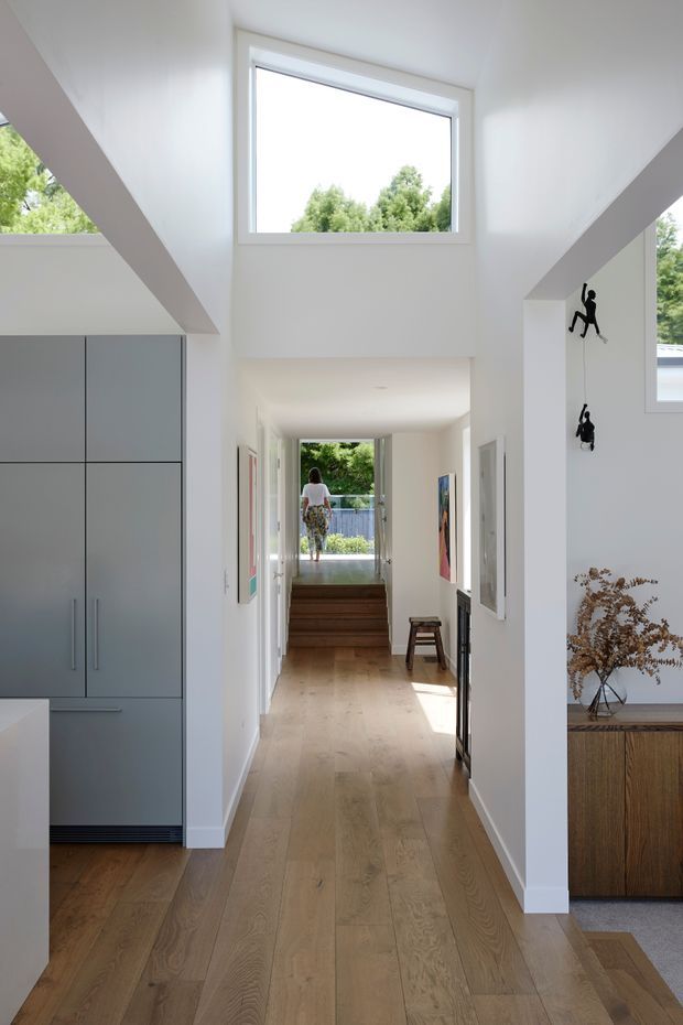Bright contemporary residential hallway or entry corridor with a warm timber floor, white walls, and a tall central void capped by a large horizontal window. The corridor leads up a short set of timber stairs toward an exterior doorway. Built-in grey cabinetry occupies the left side, while a timber console with a vase and wall-mounted decorative figures appears on the right. Natural greenery is visible through the glazing.