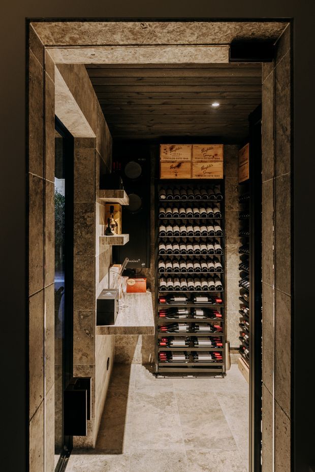 A narrow, dark, highly finished wine storage room viewed through a stone-framed doorway. The space features floor-to-ceiling wine racks filled with bottles, timber-lined ceiling boards, dark cabinetry, stone shelving and counters, and large-format pale stone flooring. Warm recessed lighting creates an intimate, luxurious atmosphere.