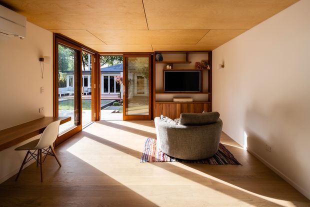 A warm, compact contemporary living room with a built-in timber media wall, television, low cabinetry and open shelving. A rounded upholstered lounge chair sits on a patterned rug facing the media unit. Floor-to-ceiling timber-framed glazing and glazed doors open toward an outdoor courtyard, while a small built-in timber desk occupies the left side. The room has a pale timber-lined ceiling, light walls and timber flooring, with strong afternoon sunlight creating long shadows.