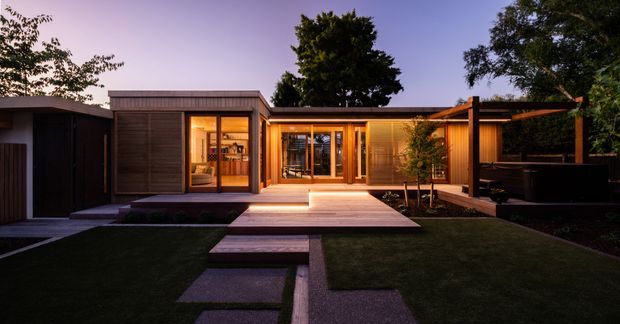 Twilight exterior view of a low-profile contemporary residential pavilion opening onto a landscaped courtyard. The composition features dark timber-clad volumes, warm timber-framed glazing, a broad timber deck and stepping-stone path across a dark lawn. A covered outdoor structure or pergola extends to the right, with warm interior lighting creating an inviting glow.