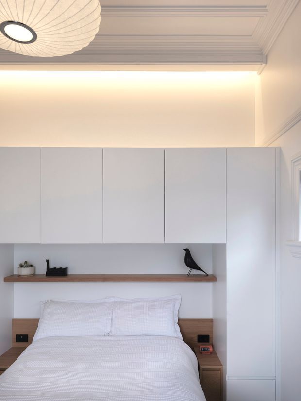 A refined bedroom with a built-in white cabinetry wall surrounding the bed, open timber display shelf, integrated bedside tables, and a tall wardrobe. The room features crisp white bedding, decorative wall and ceiling mouldings, and a sculptural pendant light, creating a calm, minimalist yet classically detailed interior.
