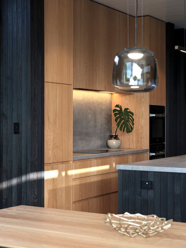 A refined contemporary kitchen with full-height warm timber cabinetry, dark vertical timber wall panelling, integrated black appliances, a grey stone splashback and benchtop, and a dark island with a light stone surface. A sculptural glass pendant hangs above the island, while sunlight creates warm highlights across the joinery and timber dining surface.