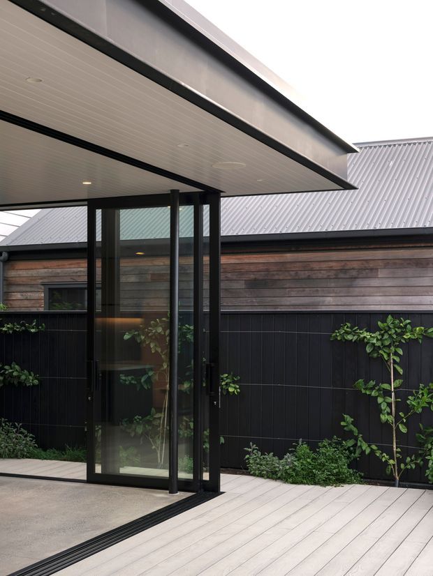 A contemporary covered entry or outdoor transition space with a black-framed glazed pivot door, pale decking, and a broad flat-roof canopy. Behind it is a single-storey building with horizontal timber cladding, dark vertical-panel fencing, a corrugated metal roof, and planted garden beds.