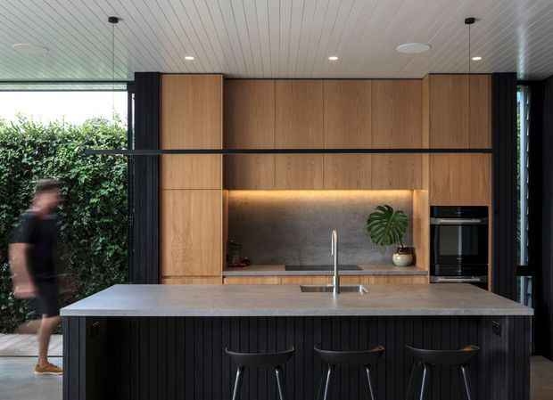 A contemporary kitchen with a large central island and three stools, full-height timber cabinetry, a dark textured splashback with warm integrated lighting, and built-in appliances. Black vertical fluted detailing frames the cabinetry and island, while large glazed openings connect the kitchen to a leafy outdoor courtyard.