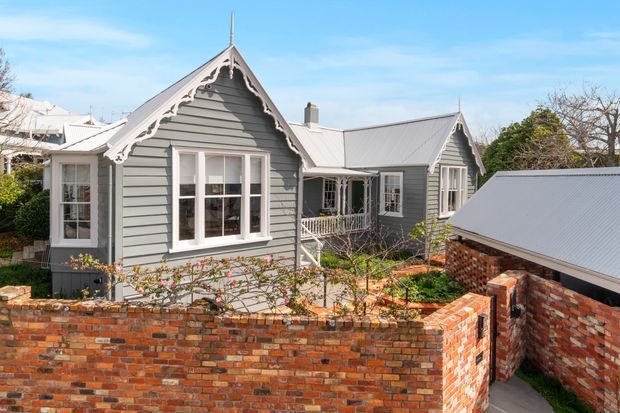 Exterior view of a charming single-storey grey weatherboard house with steep gabled roofs, decorative carved gable trim, white-framed windows, and corrugated metal roofing. A red brick garden wall and paved courtyard foreground the property, with landscaped planting around the home.