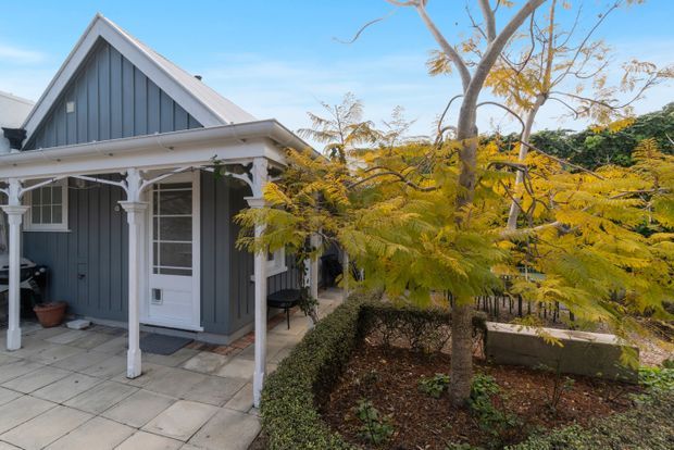Exterior view of a compact single-storey gabled home with blue-grey vertical weatherboard cladding, white trim, and a small covered verandah supported by decorative posts. A paved entry path and landscaped garden sit in front of the house.