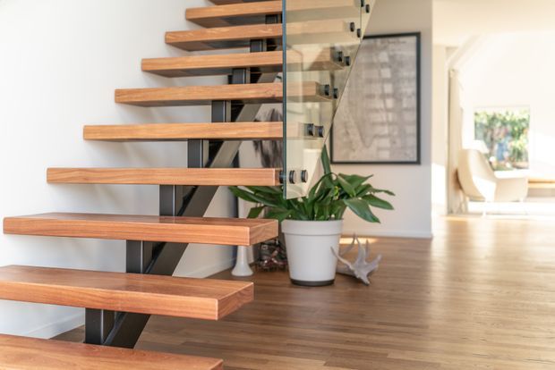 Bright contemporary interior view focused on a floating staircase with warm timber treads, dark structural supports, and a clear glass balustrade. The staircase opens onto a timber-floored living or circulation area with a potted plant and framed artwork, while a bright adjoining room is visible in the background.