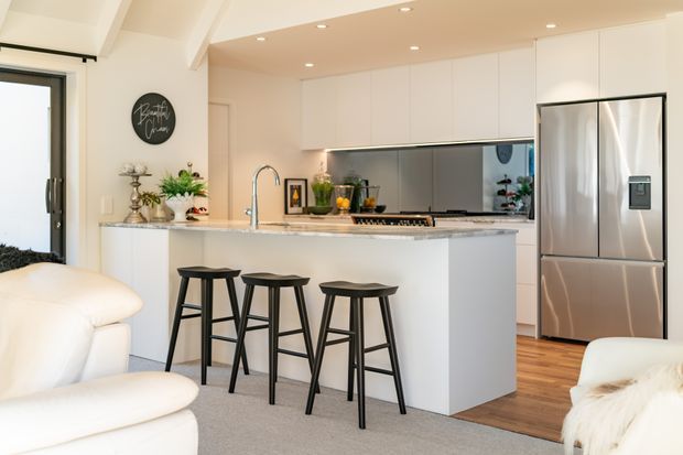 Bright contemporary open-plan kitchen with a large white island and waterfall-style benchtop, three black bar stools, white flat-panel cabinetry, a reflective dark splashback, stainless-steel French-door refrigerator, timber flooring, and recessed ceiling lighting. Part of a living area is visible in the foreground.