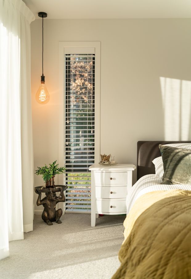A warmly lit bedroom corner featuring a partially visible bed with a dark upholstered headboard, olive-green and cream bedding, a white bedside chest, and a small decorative side table with a plant. Full-height sheer curtains and a narrow window with white plantation shutters allow strong natural sunlight into the softly neutral room. A single exposed-bulb pendant adds a warm accent.