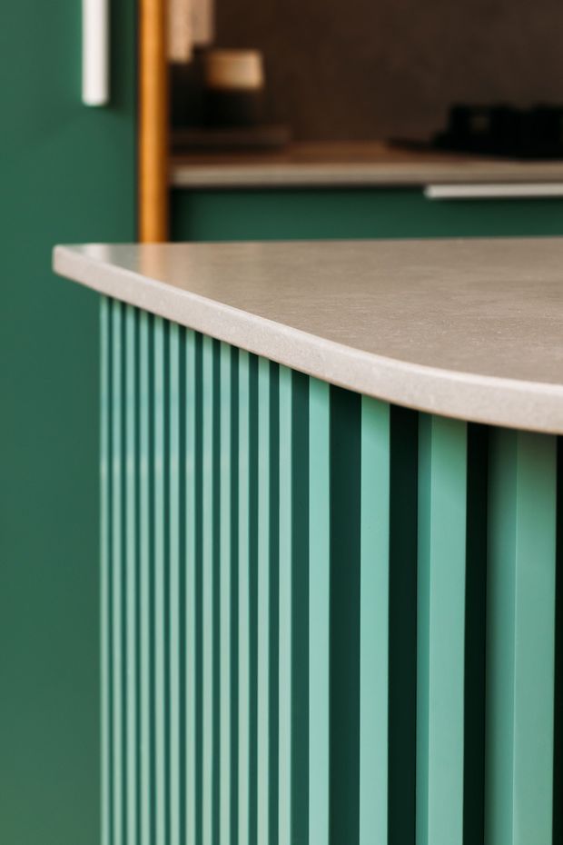 Close-up architectural detail of a curved kitchen island or counter with a pale stone-look benchtop and striking vertically ribbed green cabinetry. A dark green wall and timber-framed opening are visible in the background, with warm, directional lighting emphasizing the fluted joinery and curved edge.