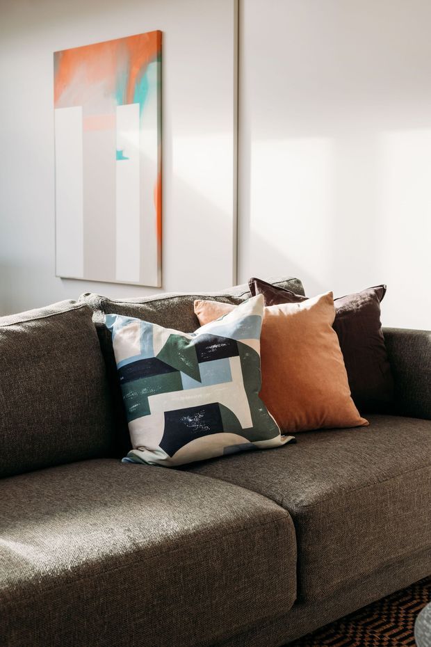 Close-up interior photograph of a dark charcoal-brown sectional sofa in a bright living room. The sofa is styled with a geometric multicoloured cushion and a warm terracotta cushion, with a large abstract artwork in orange, teal, white, and grey mounted on a pale wall behind it. Strong natural sunlight creates a warm, inviting atmosphere.