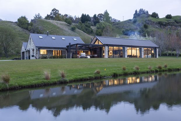 A contemporary single-storey rural residence viewed from across a landscaped pond. The long, low building combines dark metal pitched roofs, extensive glazing, dark structural framing, and a stone-clad gabled wing. Warm interior lighting is visible through the windows, while rolling grassy hills and mature trees form the background.