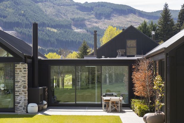 A contemporary alpine courtyard viewed from outdoors, framed by dark-clad pitched-roof buildings. The central glazed pavilion has expansive floor-to-ceiling sliding glass doors, connecting the paved courtyard to a green lawn and mountain setting beyond. A stone masonry wall, black metal chimney, outdoor seating and dining furniture, planters, and clipped landscaping complete the scene.