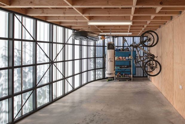 A spacious industrial-style garage or multipurpose storage area with a polished concrete floor, exposed timber ceiling joists, plywood wall lining, and expansive black-framed glazed panels. Bicycles are mounted on the wall, with shelving and sporting equipment at the far end. The open structure and abundant glazing create a bright, utilitarian workspace.