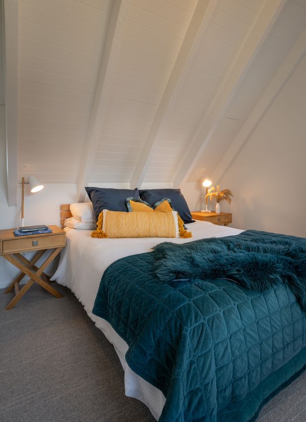 A cosy attic-style bedroom with a sloped white timber-lined ceiling and exposed beams. The room features a neatly made bed with layered white bedding, navy and mustard cushions, and a deep teal quilted throw. Matching bedside tables, warm bedside lamps, a small decorative vase, and neutral carpeting create a comfortable, refined atmosphere.