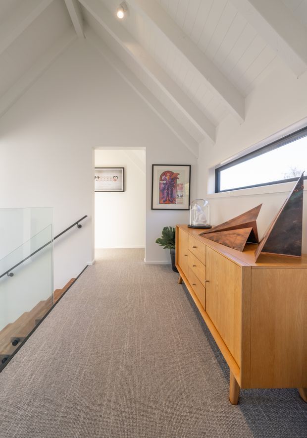 A bright upper-level hallway or landing with a pitched, exposed-beam white ceiling, a large horizontal window, glass stair balustrade, and carpeted flooring. A warm timber sideboard with drawers occupies the right side, topped with sculptural artwork and a decorative glass object. Framed artwork and a plant add visual interest, while a doorway leads to another area in the background.